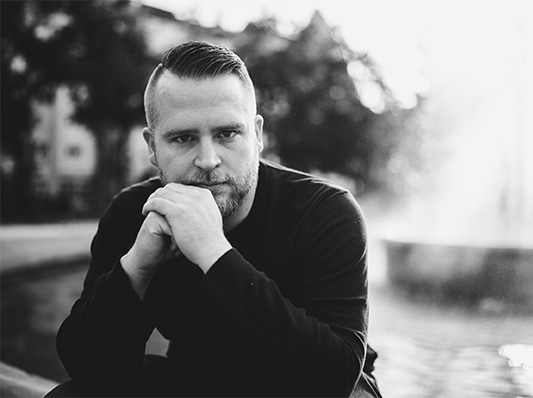 Ein Mann mit Bart sitzt nachdenklich auf dem Rand eines Springbrunnens in einem Park, die Hände vor dem Mund verschränkt.