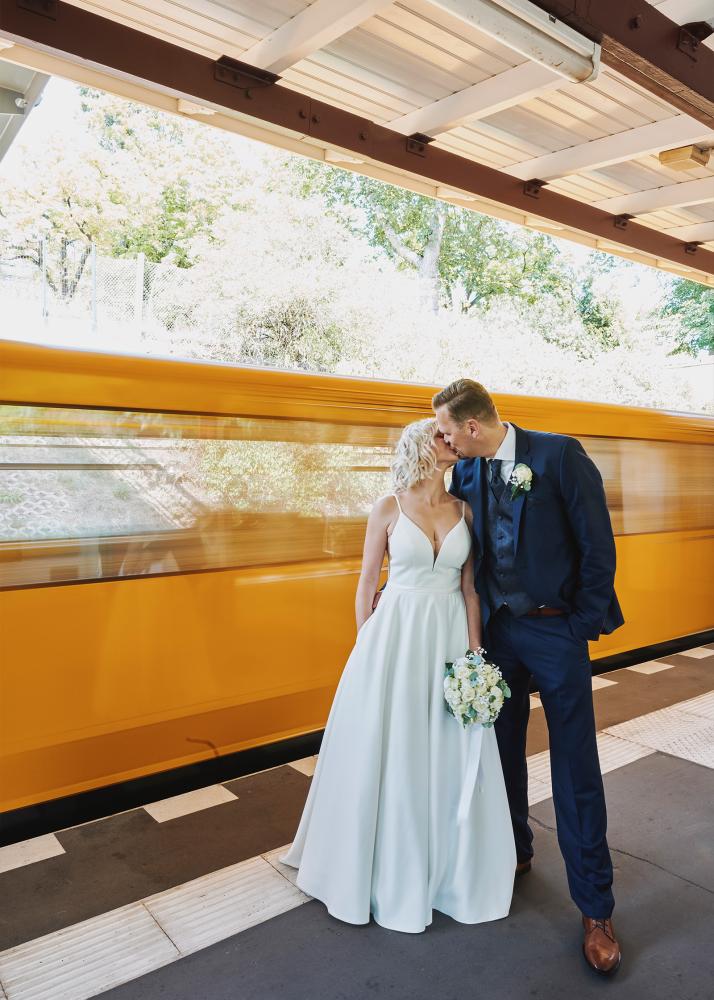 Ein Brautpaar auf einem Bahnsteig, das sich küsst, während ein Zug vorbeifährt.