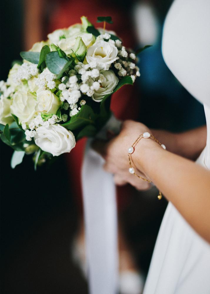 Eine Hand hält einen Brautstrauß mit weißen Rosen und Eukalyptusblättern, während ein Arm mit einem Armband zu sehen ist.