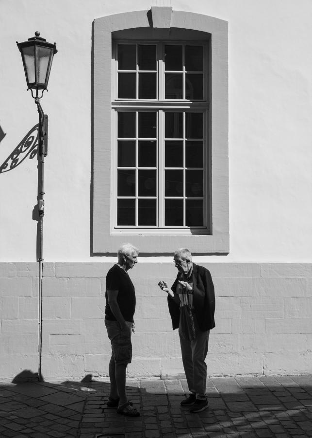 Zwei Männer unterhalten sich intensiv vor der hellen, sonnenbeschienenen Fassade eines Hauses. Mit Licht- und Schattenspiel in Schwarz und Weiß.