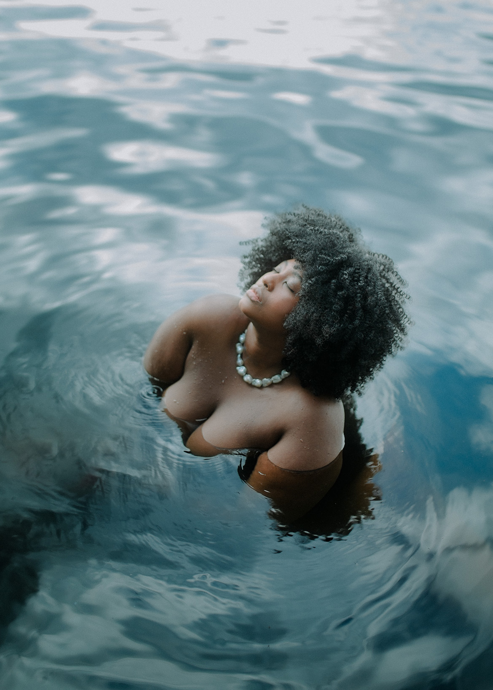 Eine Frau mit Afro steht in einem Gewässer und hält ihren Kopf mit geschlossenen Augen dem Himmel entgegen.