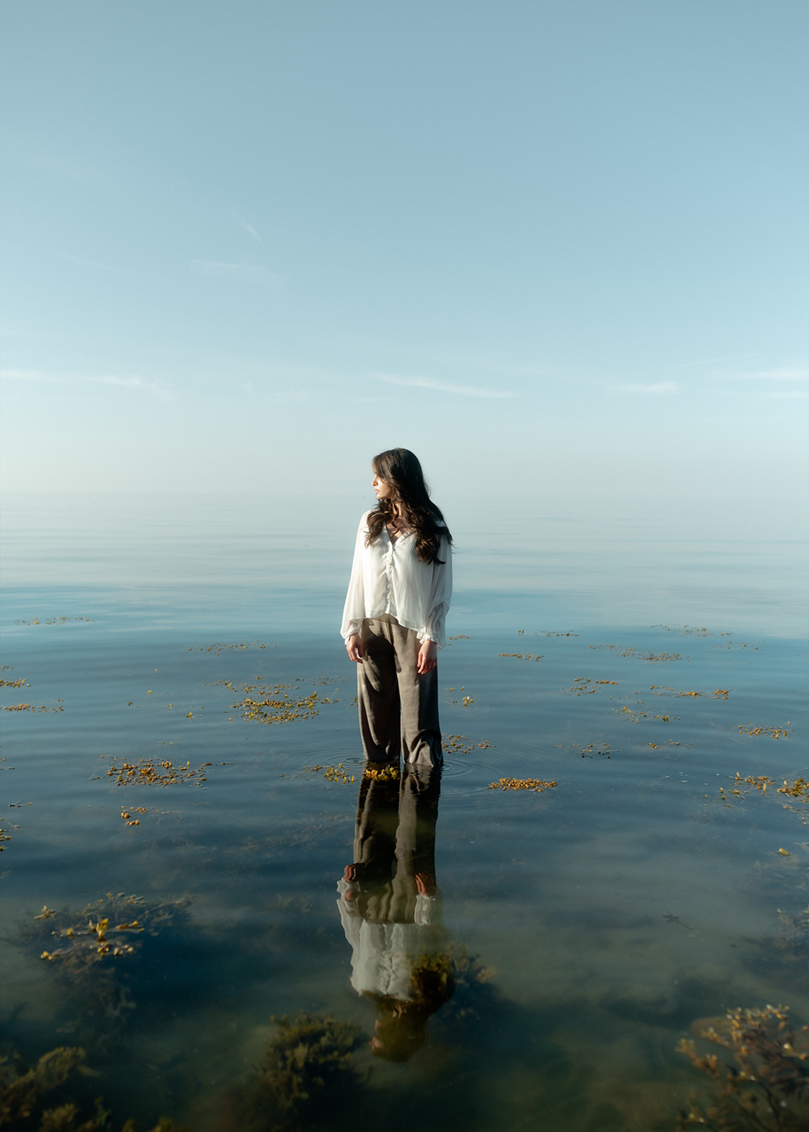 Eine Frau steht im flachen Wasser eines Sees, trägt eine weiße Bluse und graue Hosen, und ihr Spiegelbild ist im Wasser zu sehen.