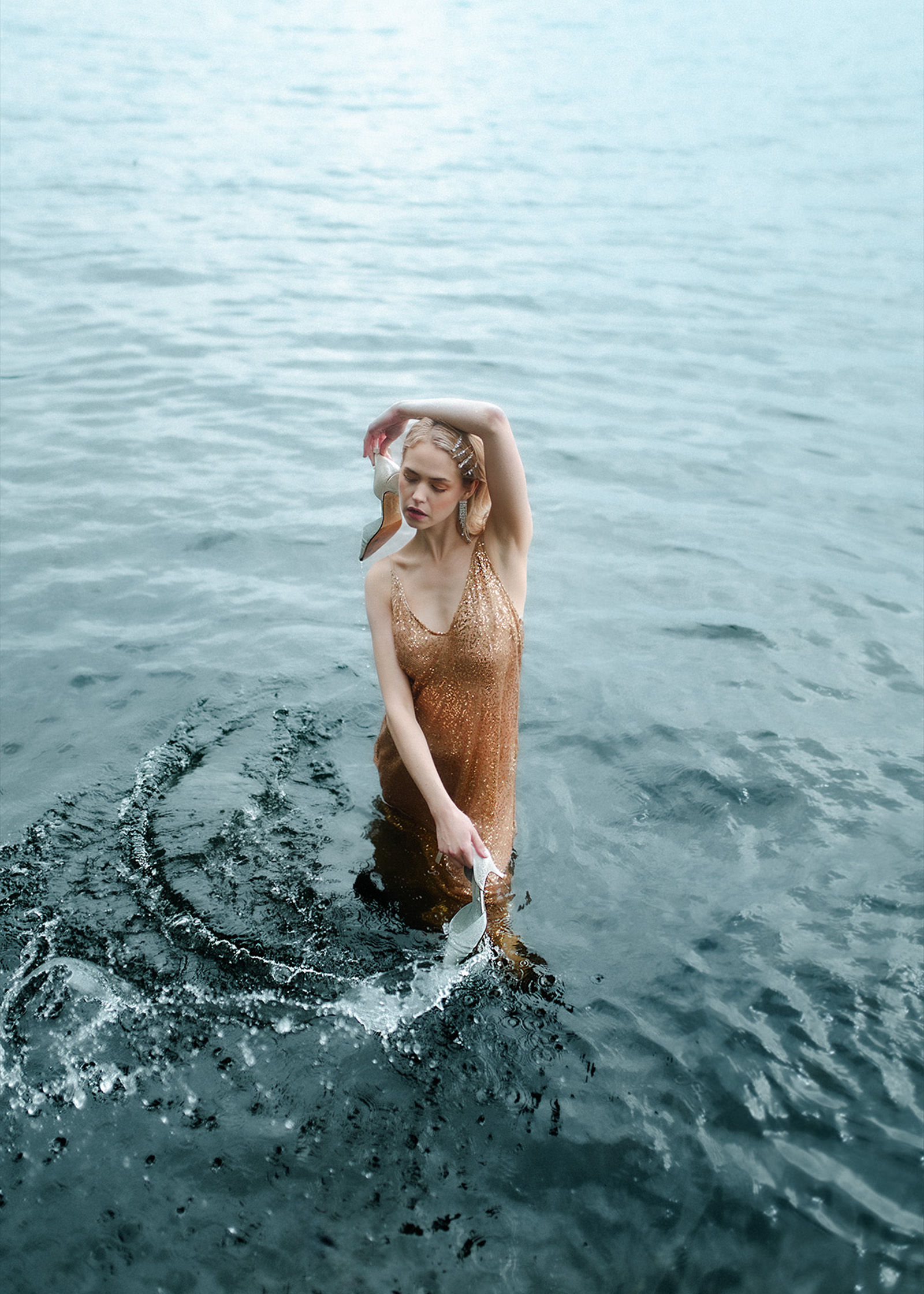 Eine Frau in einem rostbraunen Kleid steht im wasser und zieht mit einem ihrer Schuhe, welche sie in den Händen hält, einen Halbkreis aus Wellen um sich.