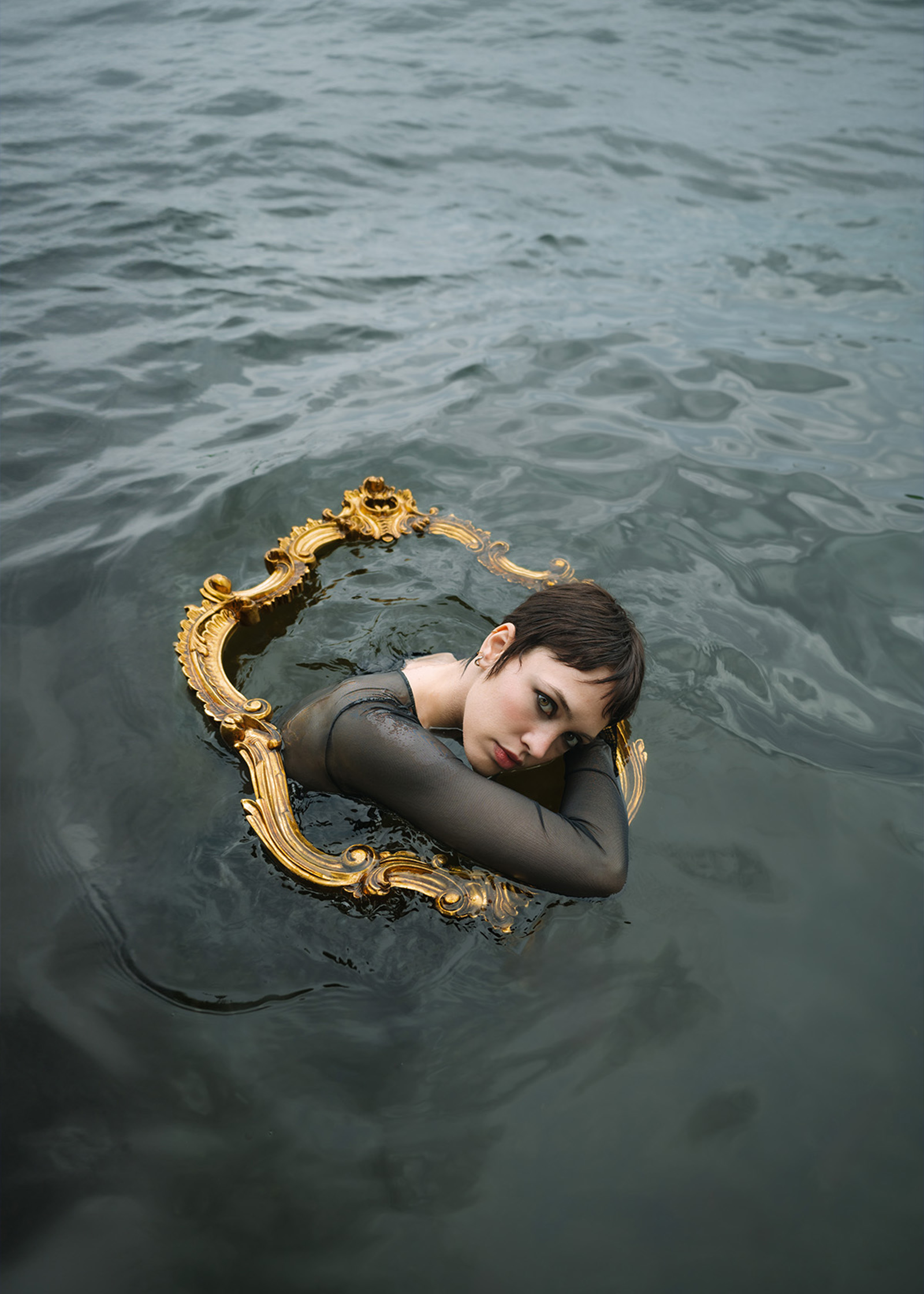 Eine Frau mit kurzen schwarzen Haaren schwimmt in einem See und schaut durch den Rahmen eines Spiegels aus dem Wasser.