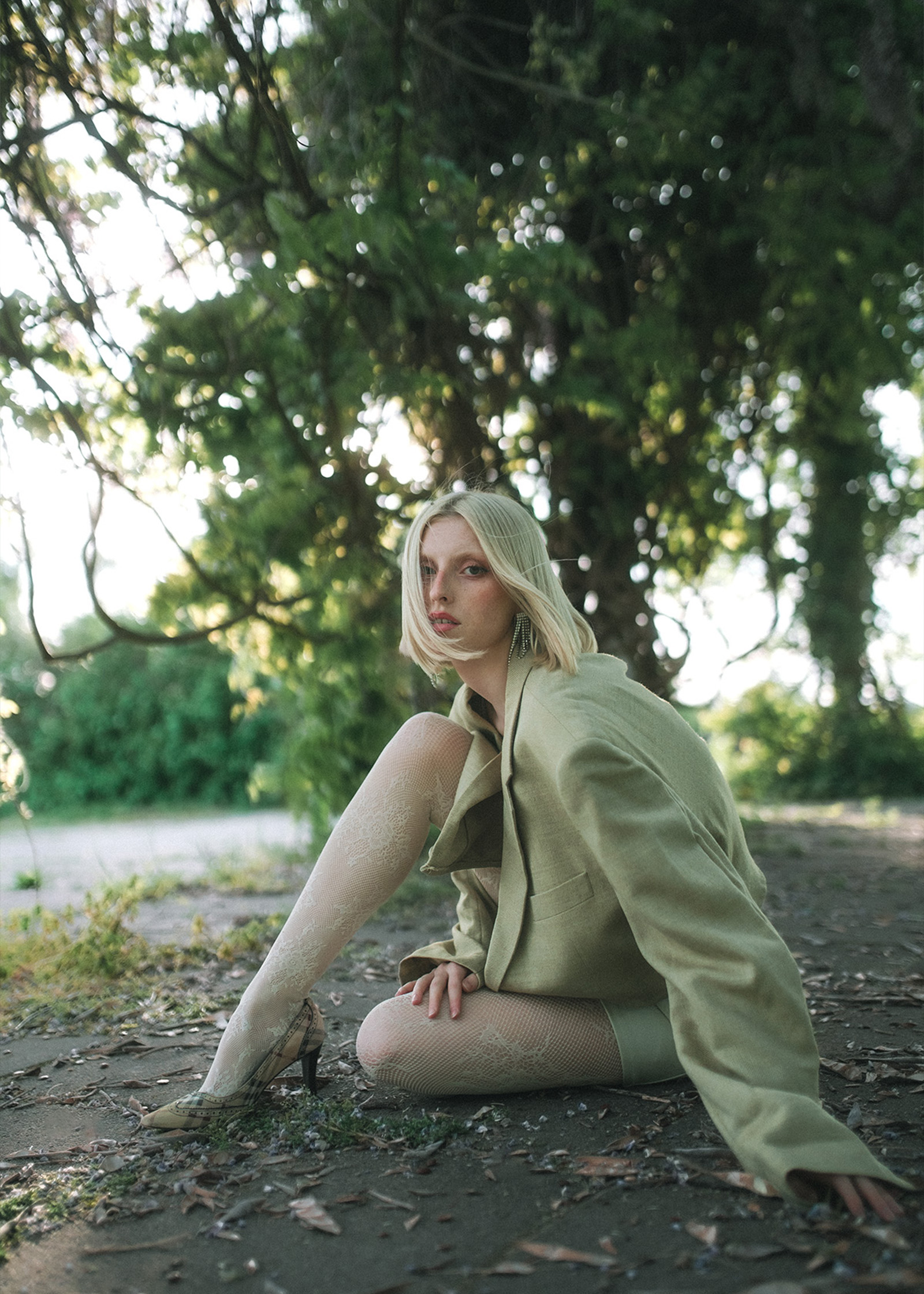 Eine Frau mit hellem Haar sitzt unter einem Baum, trägt einen grünen Blazer und ein durchsichtiges Oberteil.