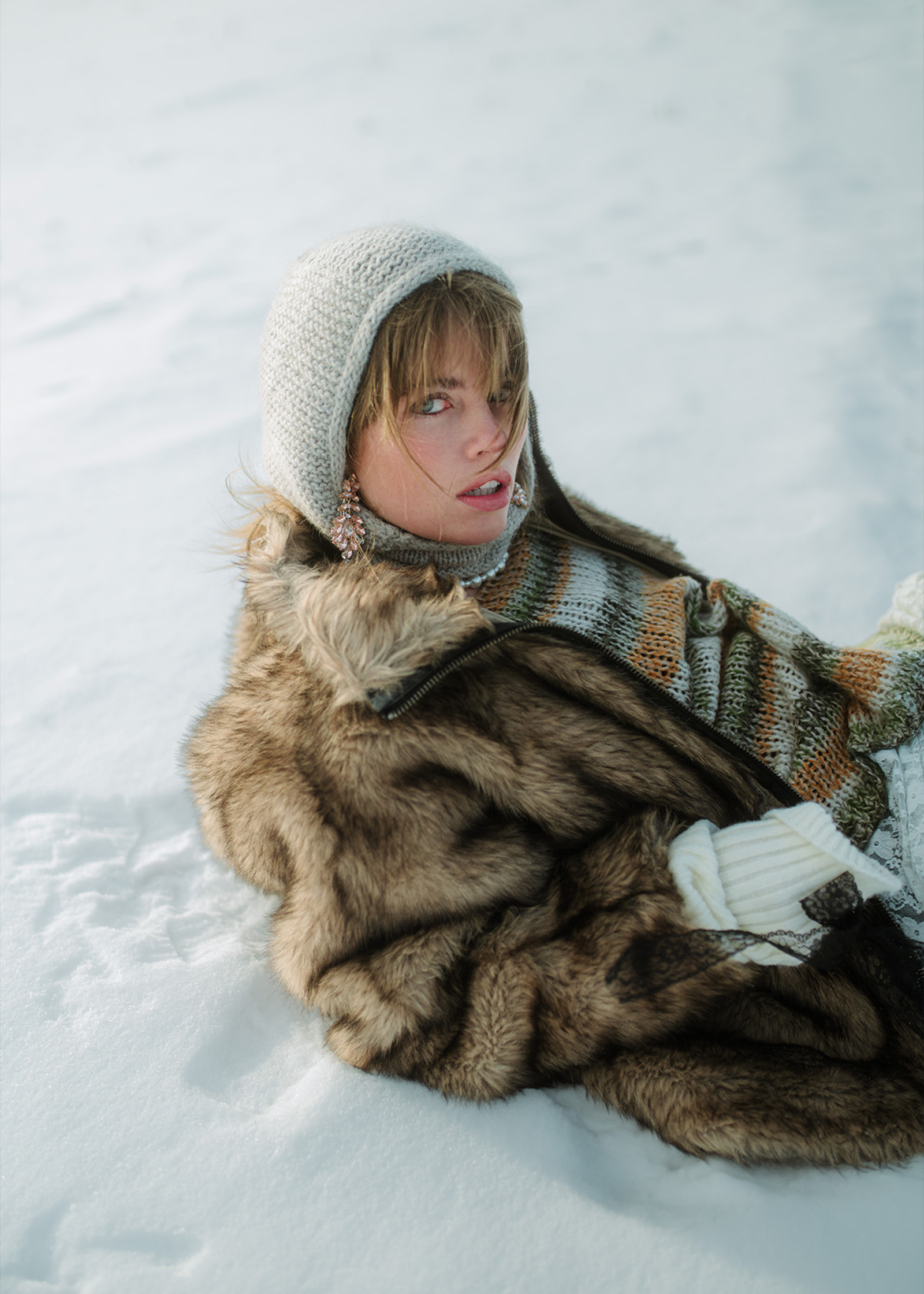 Eine Frau mit hellem Haar sitzt im Schnee, trägt einen dicken, braunen Mantel und einen grauen Schal.