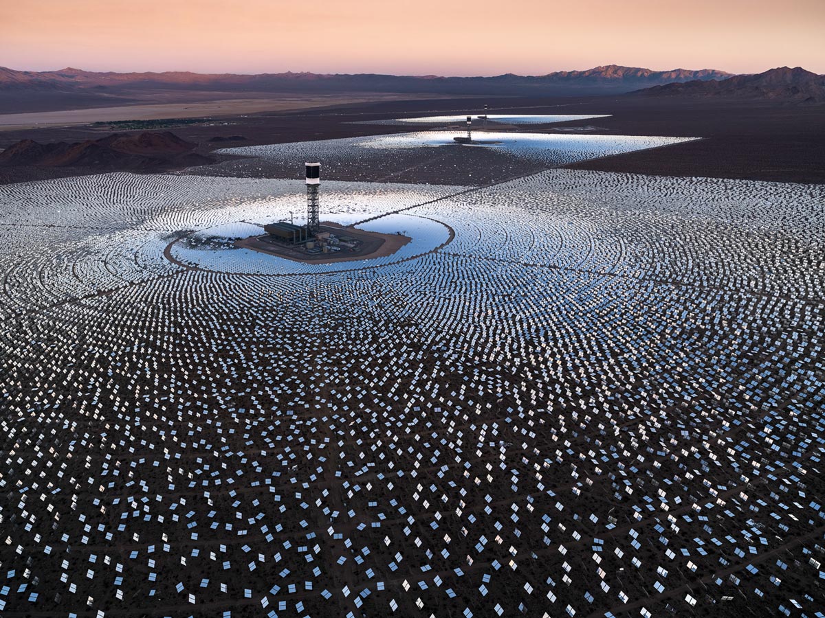Eine ausgedehnte Solaranlage in der Wüste, mit konzentrischen Kreisen von Spiegeln, die das Sonnenlicht auf zentrale Türme reflektieren, umgeben von einer weitläufigen, trockenen Landschaft.