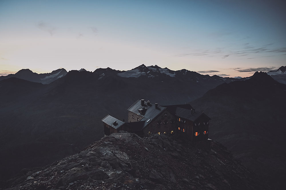 Ein belebtes Haus auf einem Berg, während im Hintergrund die letzten Strahlen der Sonne zu sehen sind, welche bereits hinter den hohen Bergen in der Ferne untergegangen ist.