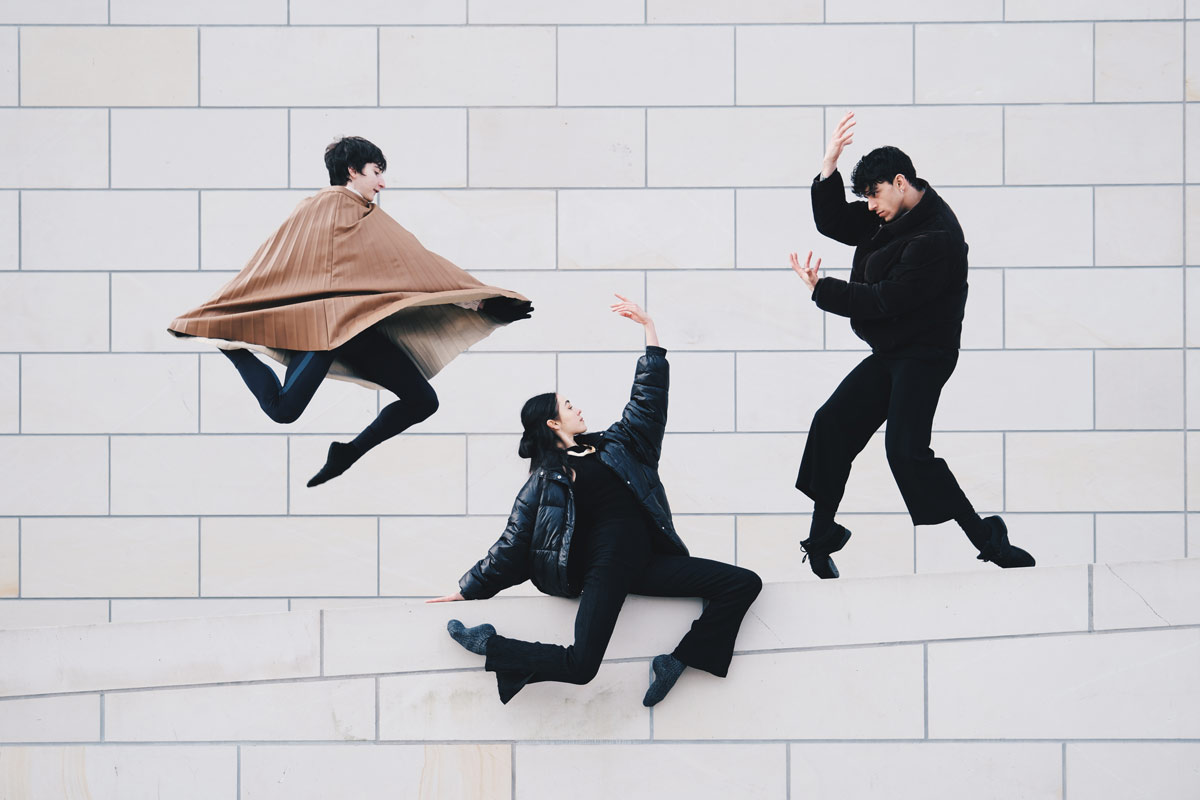 Drei Tänzer in verschiedenen Posen vor einer weißen Ziegelwand, einer in der Luft, einer auf dem Boden und einer in einer Kampfstellung.