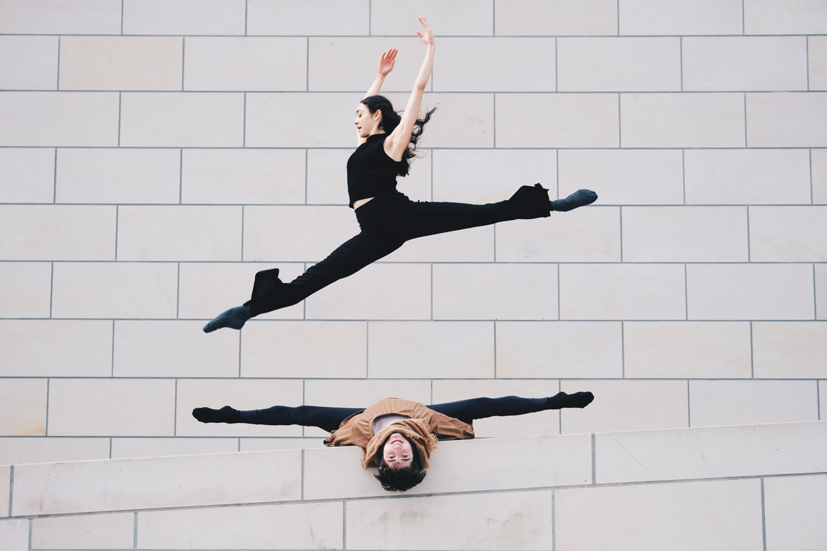 Eine Tänzerin in schwarzer Kleidung springt vor einer weißen Ziegelwand, während ein anderer Tänzer in brauner Kleidung auf dem Boden liegt.
