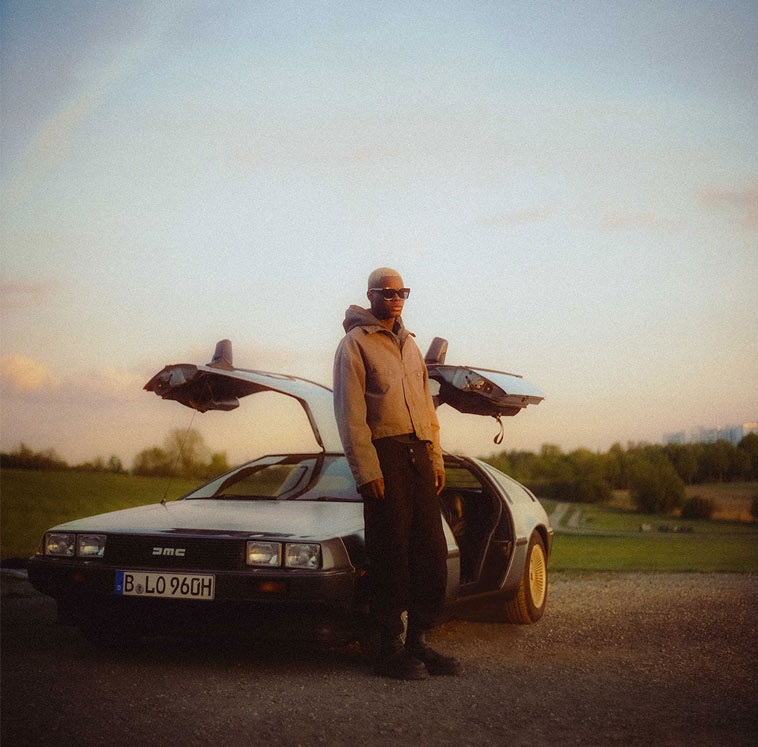 Eine Person steht vor einem DeLorean-Auto mit geöffneten Türen und schaut in die Kamera.