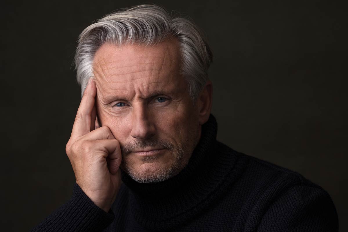 Ein älterer Mann mit grauen Haaren und einem schwarzen Rollkragenpullover hält sich die Hand an die Wange und blickt nachdenklich in die Kamera.