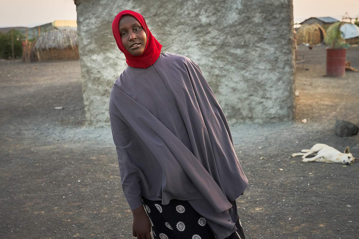 Eine Frau in einem grauen Umhang und einem roten Kopftuch steht vor einem Haus aus Stein und Stroh, mit einem Hund, der im Hintergrund liegt.