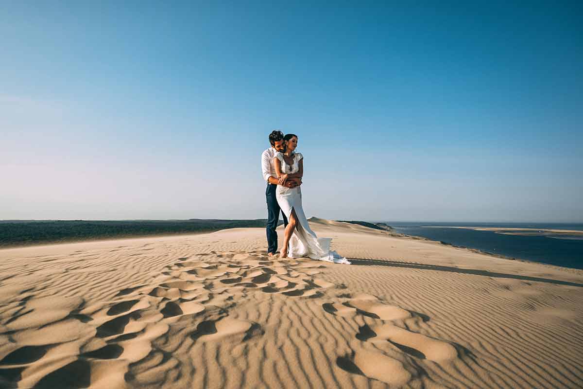 Ein Hochzeitspaar umarmt sich lachend auf einer Sanddüne direkt am Meer.