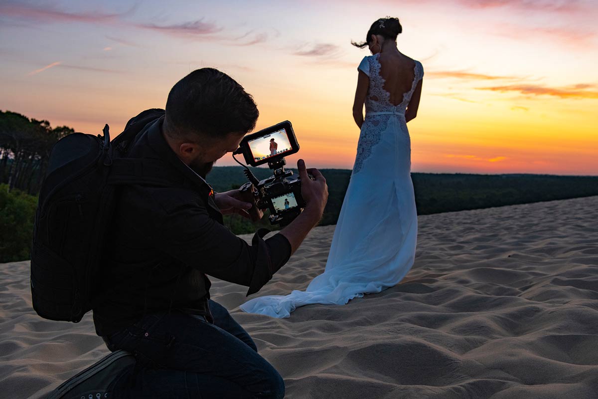 Ein Fotograf fotografiert eine Frau in einem Hochzeitskleid auf einer Sanddüne, während im Hintergrund die Sonne untergeht.