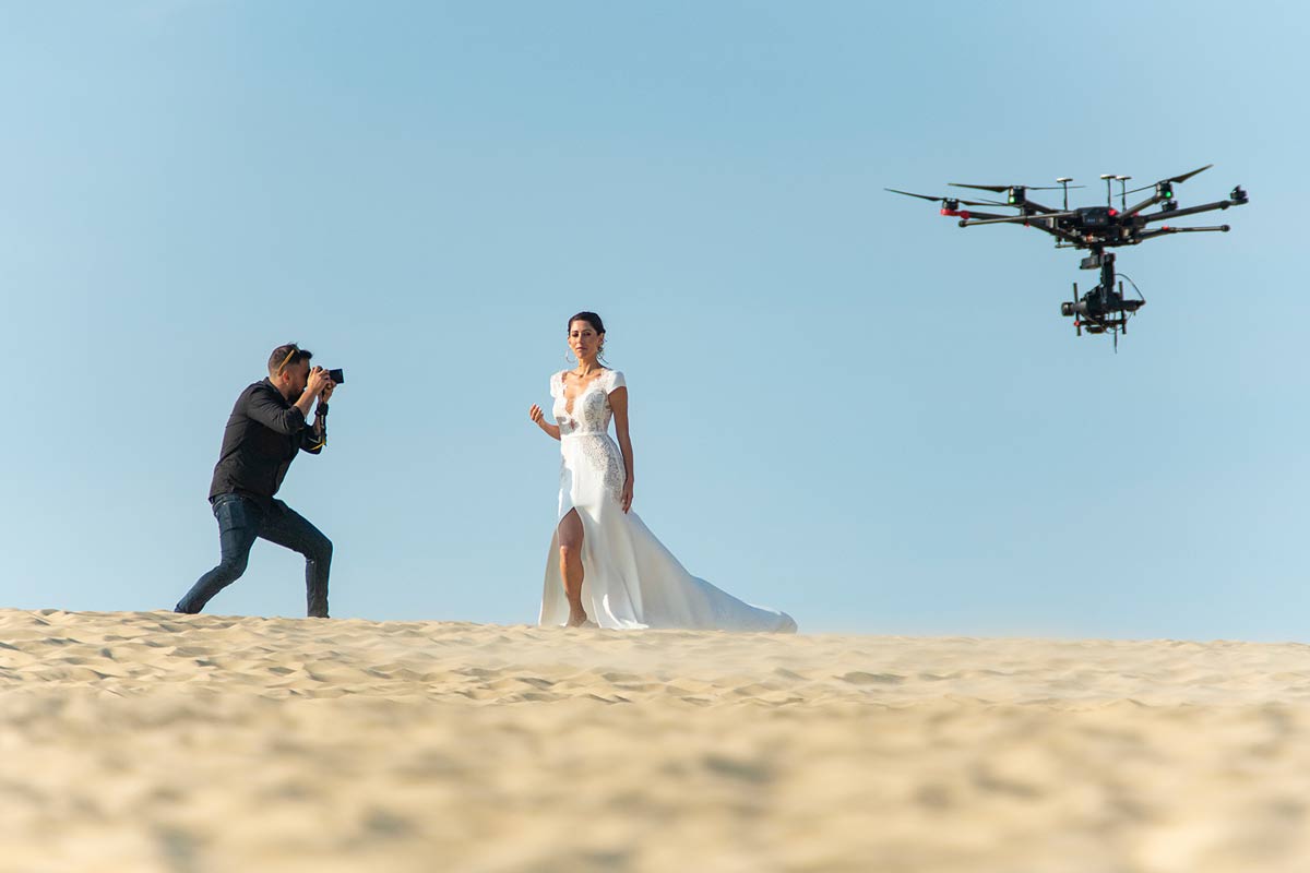 Ein Fotograf fotografiert eine Frau in einem weißen Hochzeitskleid auf einer Sanddüne. Außerdem sieht man eine fliegende Drohne.