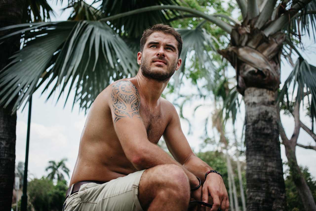 Ein Mann mit Schulter tätowierung in einer kurzen Hose sitzend unter Palmenbäumen.