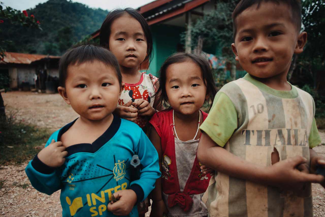 Kinder mit schmutzigen Klamotten, die unsicher in die Kamera gucken. Im Hintergrund sieht man mehrere Gebäude.