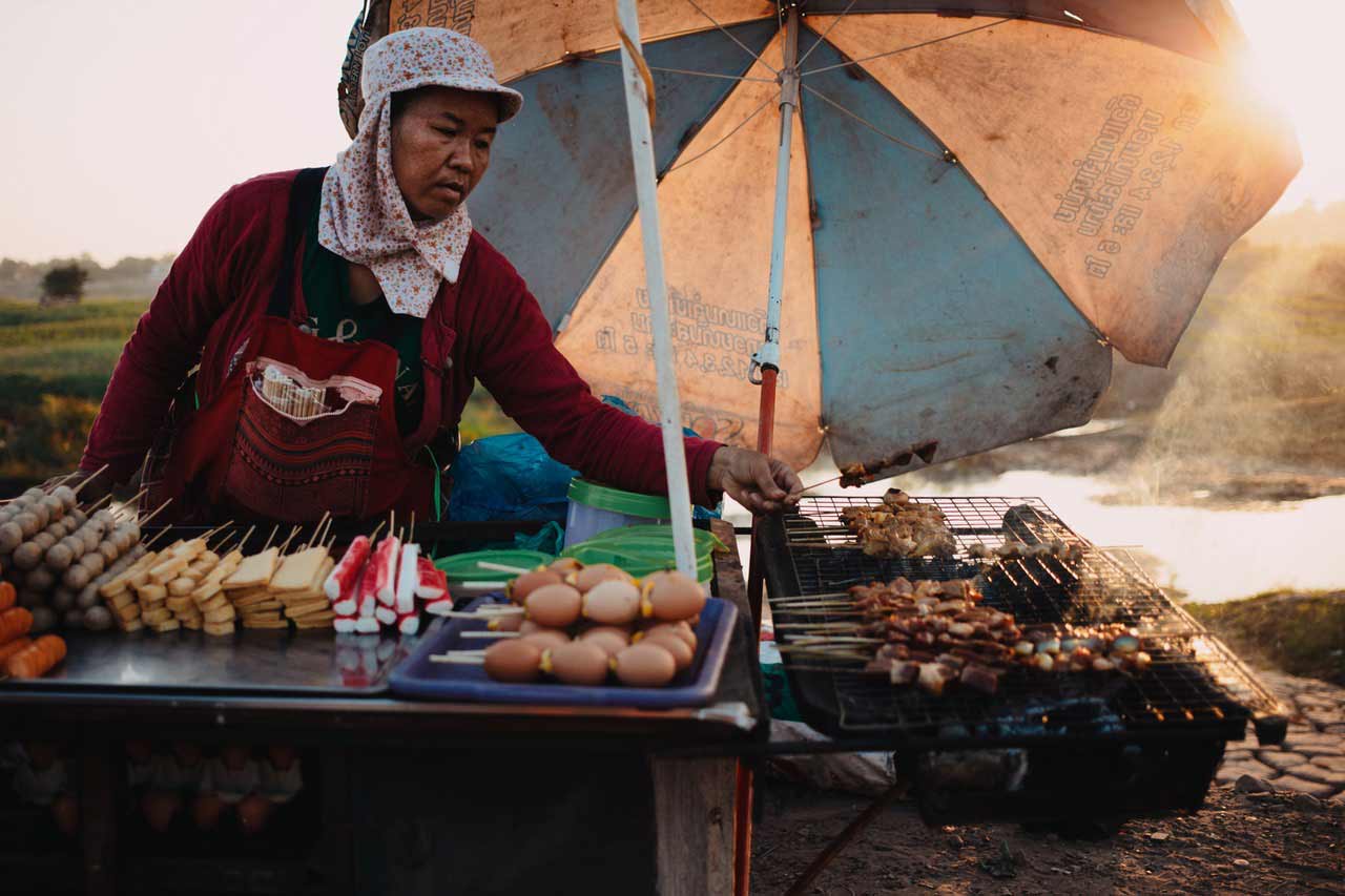 Ein kleiner, etwas heruntergekommener Straßen Imbiss mit einer Verkäuferin, welche die Grillwaren wendet.