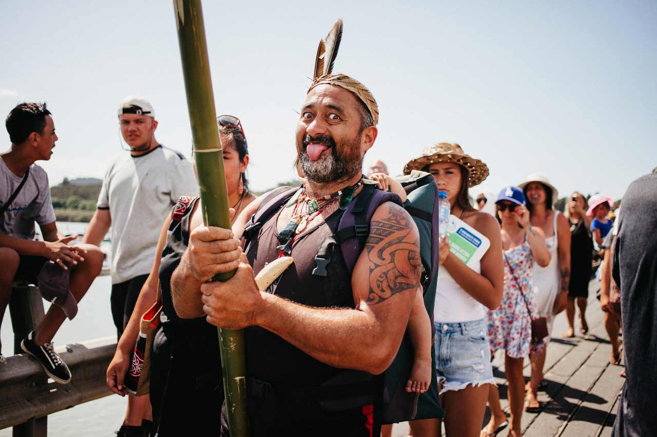 Ein Mann mit einer geflochtenen, mit Federn geschmückten Kopfbedeckung hält einen Bambusstamm und zieht eine Grimasse.