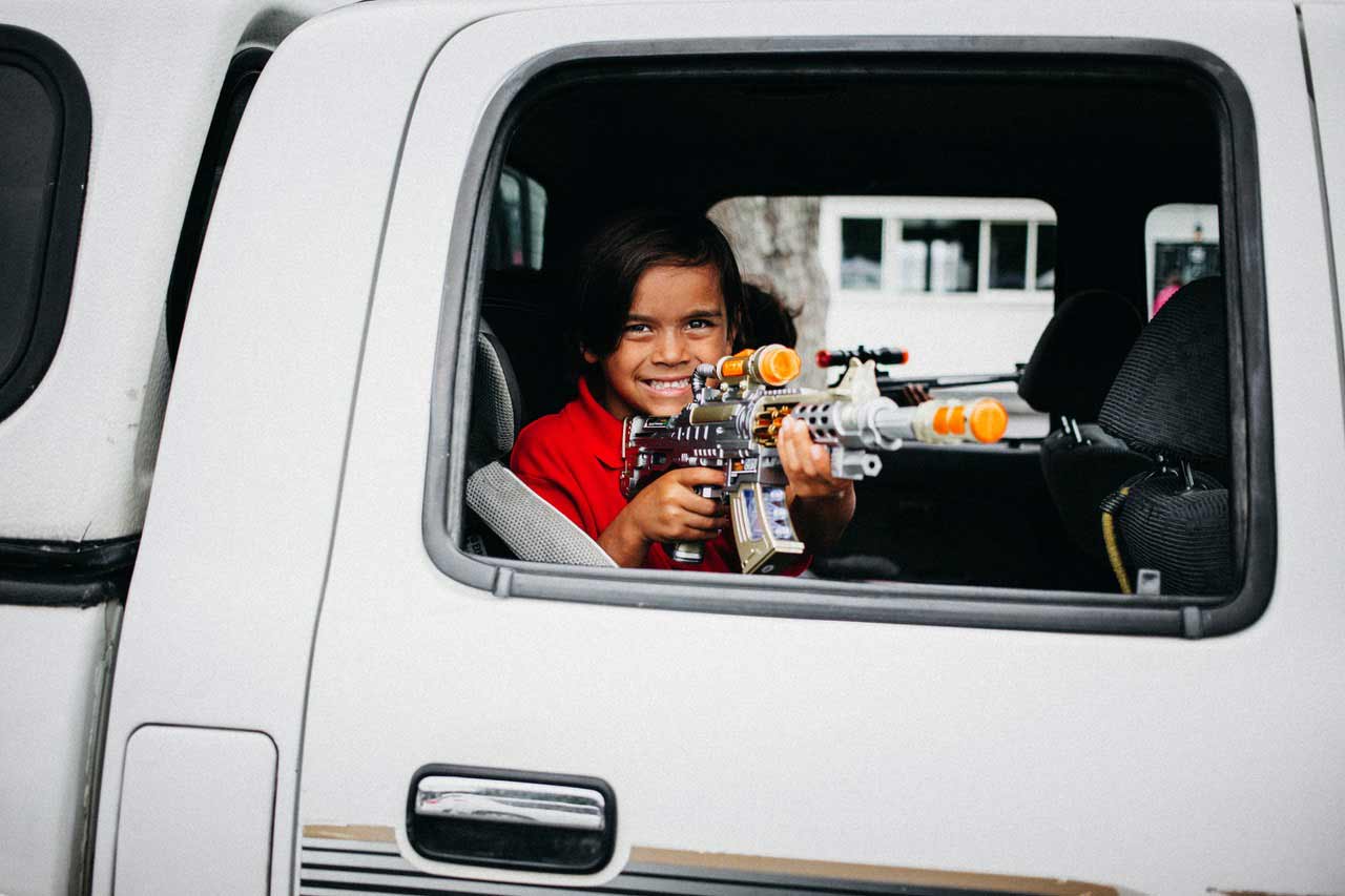 Ein Junge hält eine Spielzeugwaffe aus dem Fenster eines Autos, so als würde er sie bedienen und lacht in die Kamera.