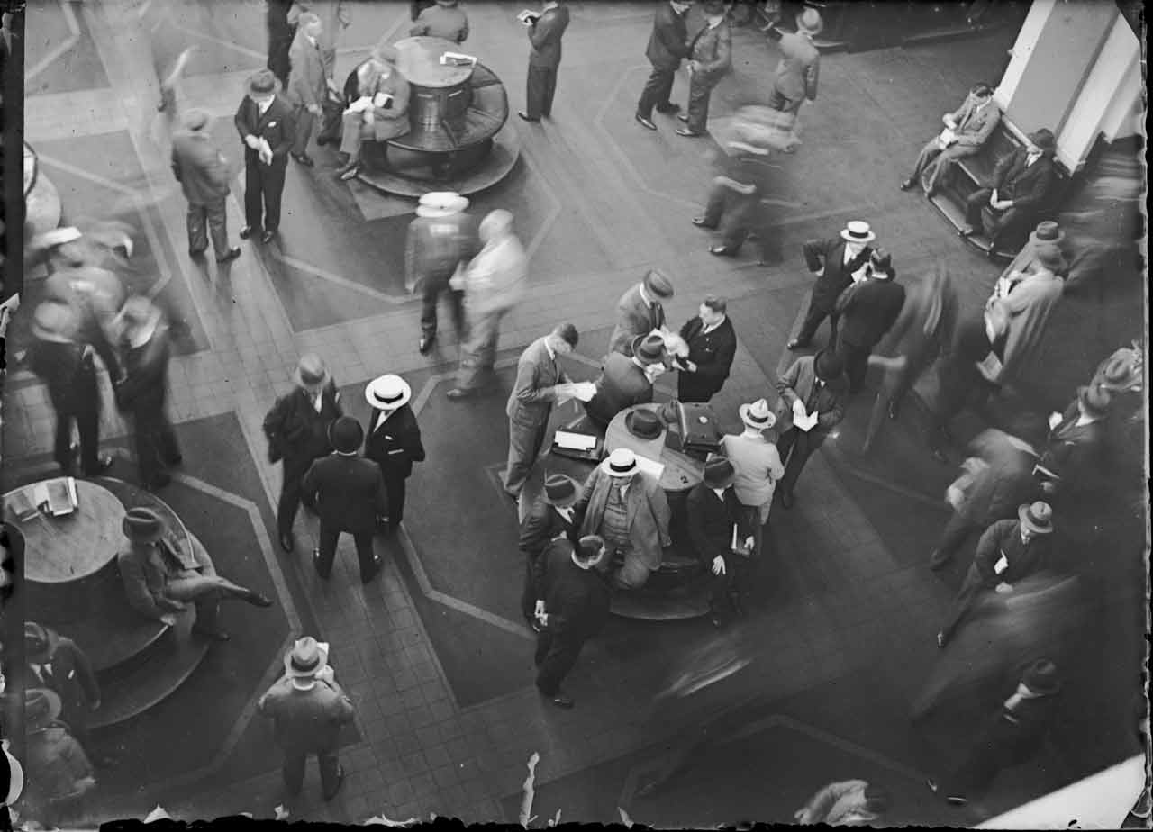 Eine Gruppe von Männern in Anzügen steht, sitzt und läuft in einem Raum mit geometrischen Bodenmustern und runden Tischen.