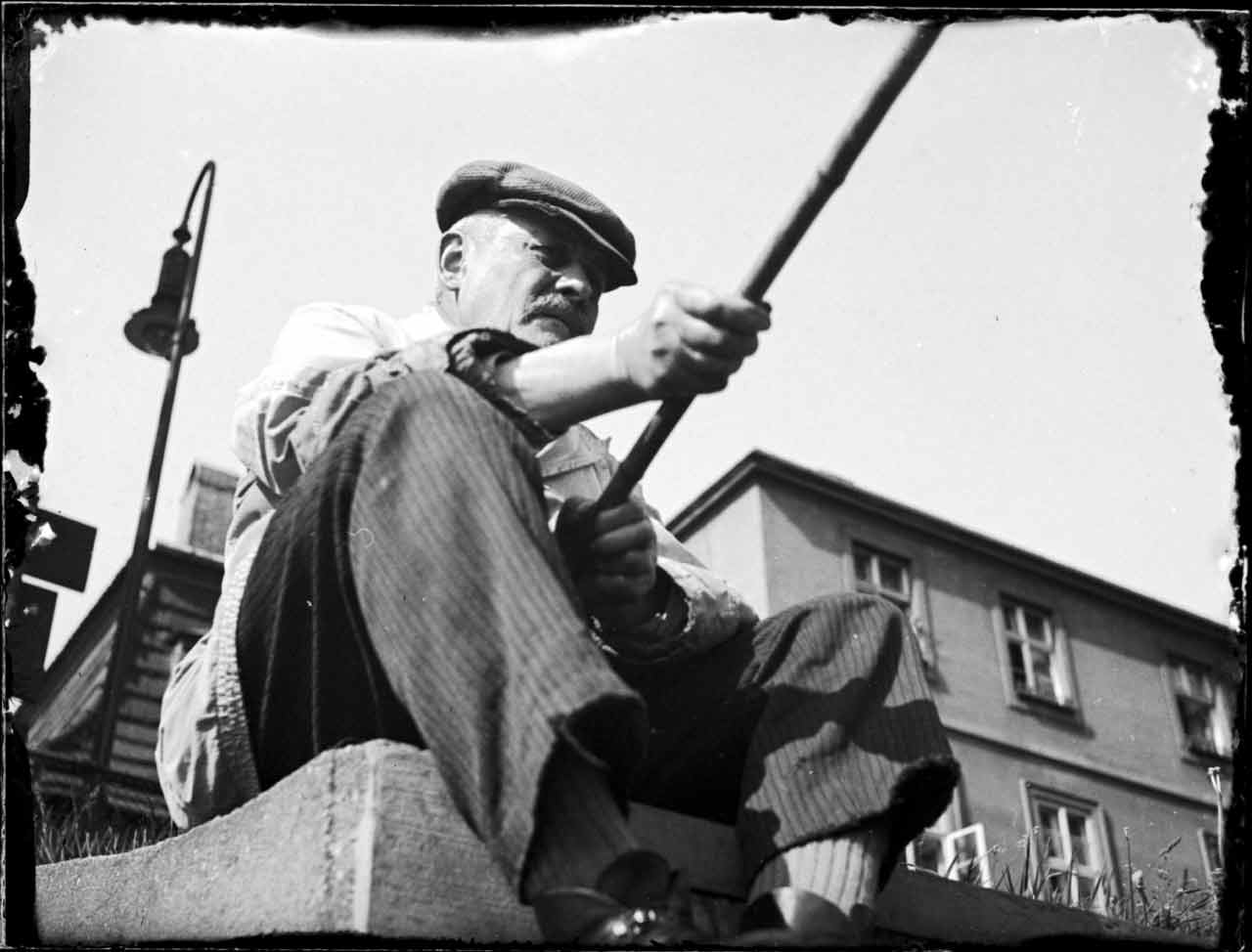 Ein Mann mit einer Kappe und einem Hemd mit aufgerollten Ärmeln hält einen Stock und sitzt auf einem Steinblock.