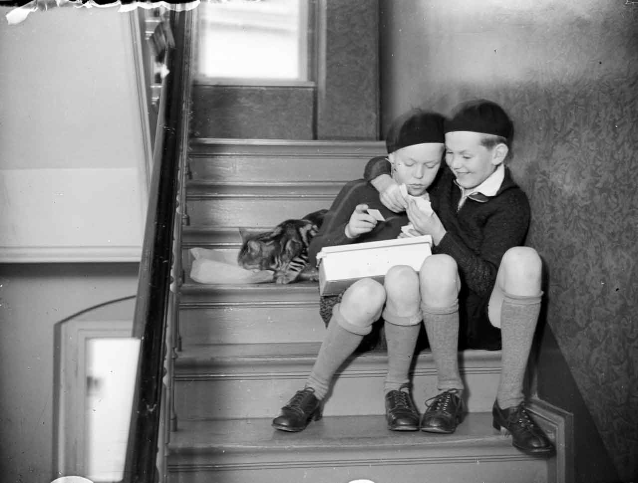 Zwei Kinder sitzen auf einer Treppe und spielen Karten, während eine Katze neben ihnen liegt.