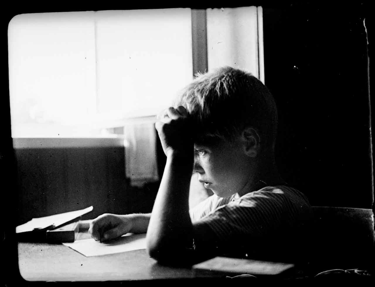 Ein Junge sitzt an einem Tisch und schreibt in ein Buch, während er sich mit der Hand an den Kopf fasst.