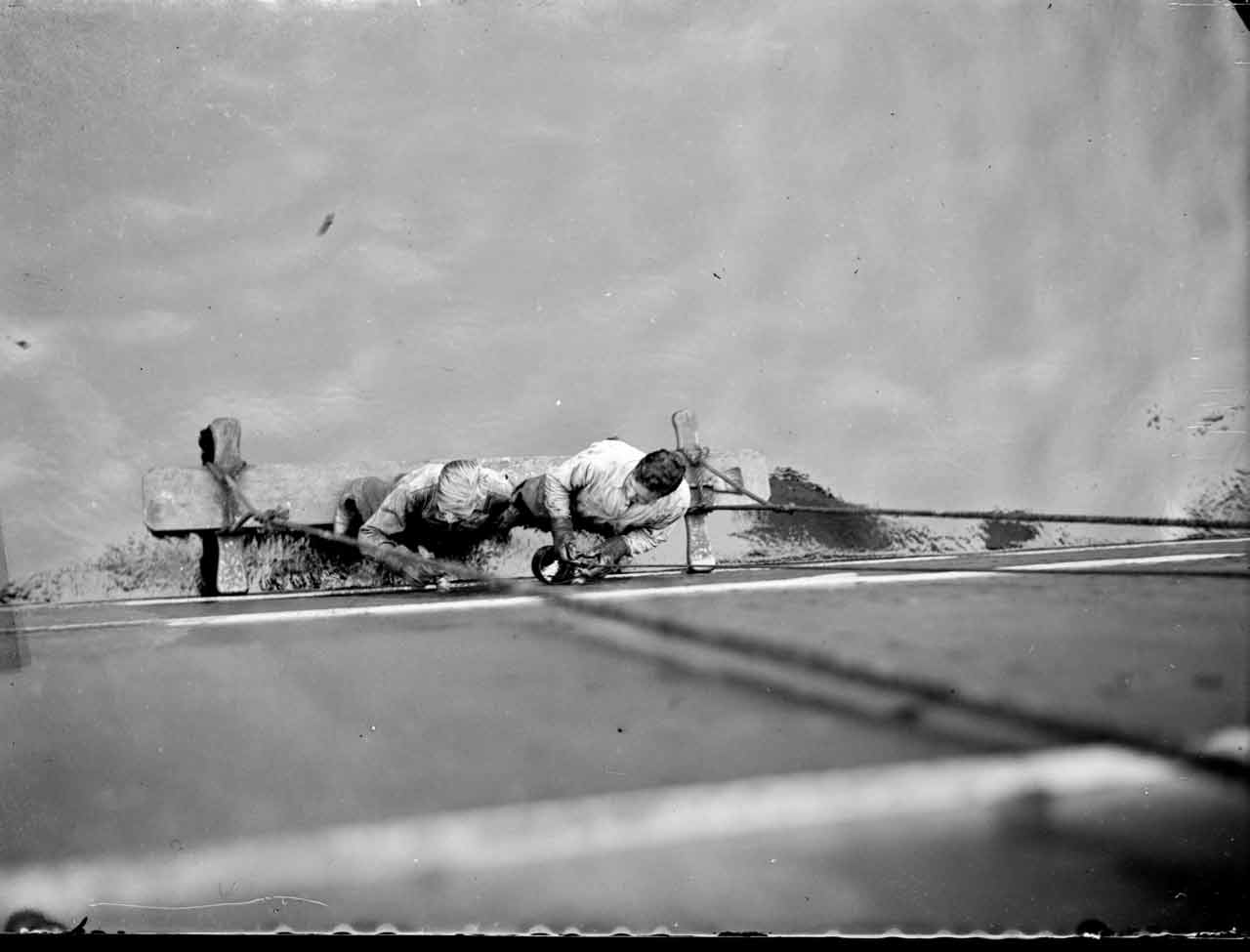 Zwei Männer liegen auf dem Boden und arbeiten an einer Wand, während sie auf einem Brett an Seilen direkt über dem Wasserspiegel stehen.