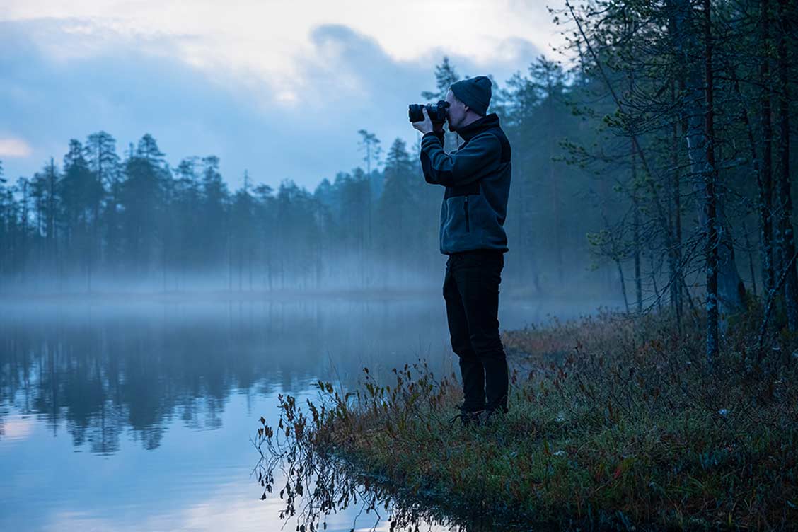 Ein Fotograf am Ufer eines Sees mit Nebelschwaden darüber.