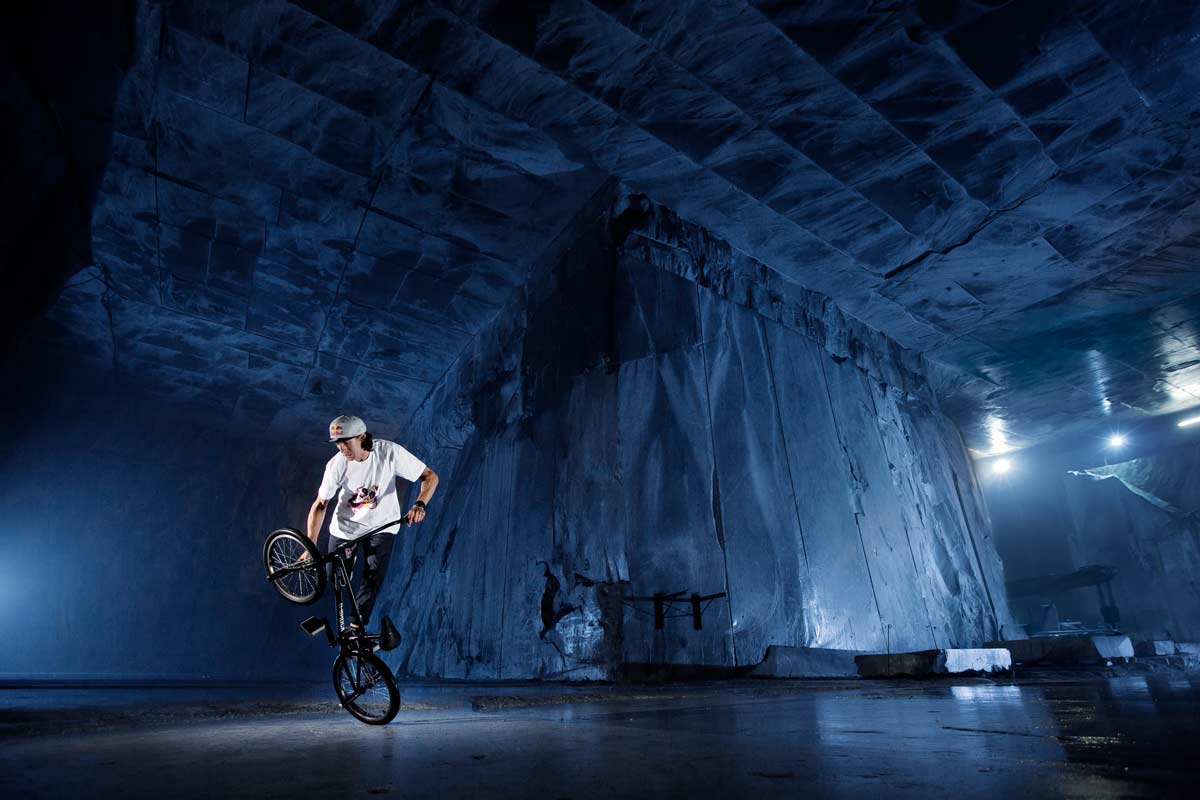 Ein Athlet macht ein BMX-Trick in einer Halle mit hohen rauen Steinwänden, die schwach beleuchtet werden.